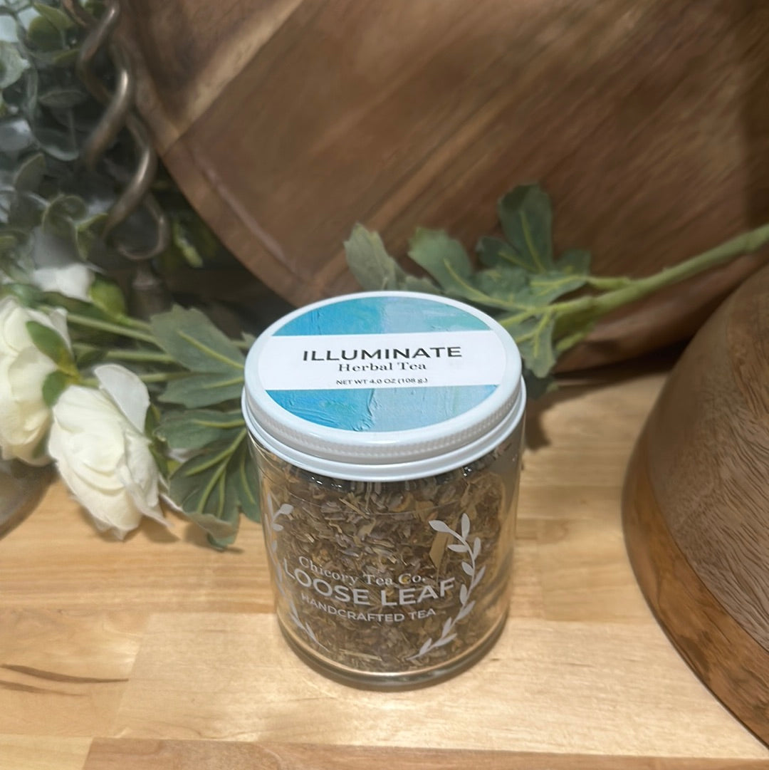 An artistic product photo of Chicory Tea Co.'s Illuminate Herbal Tea in the jar, from the front on a wood floor with flowers and barrel in the background