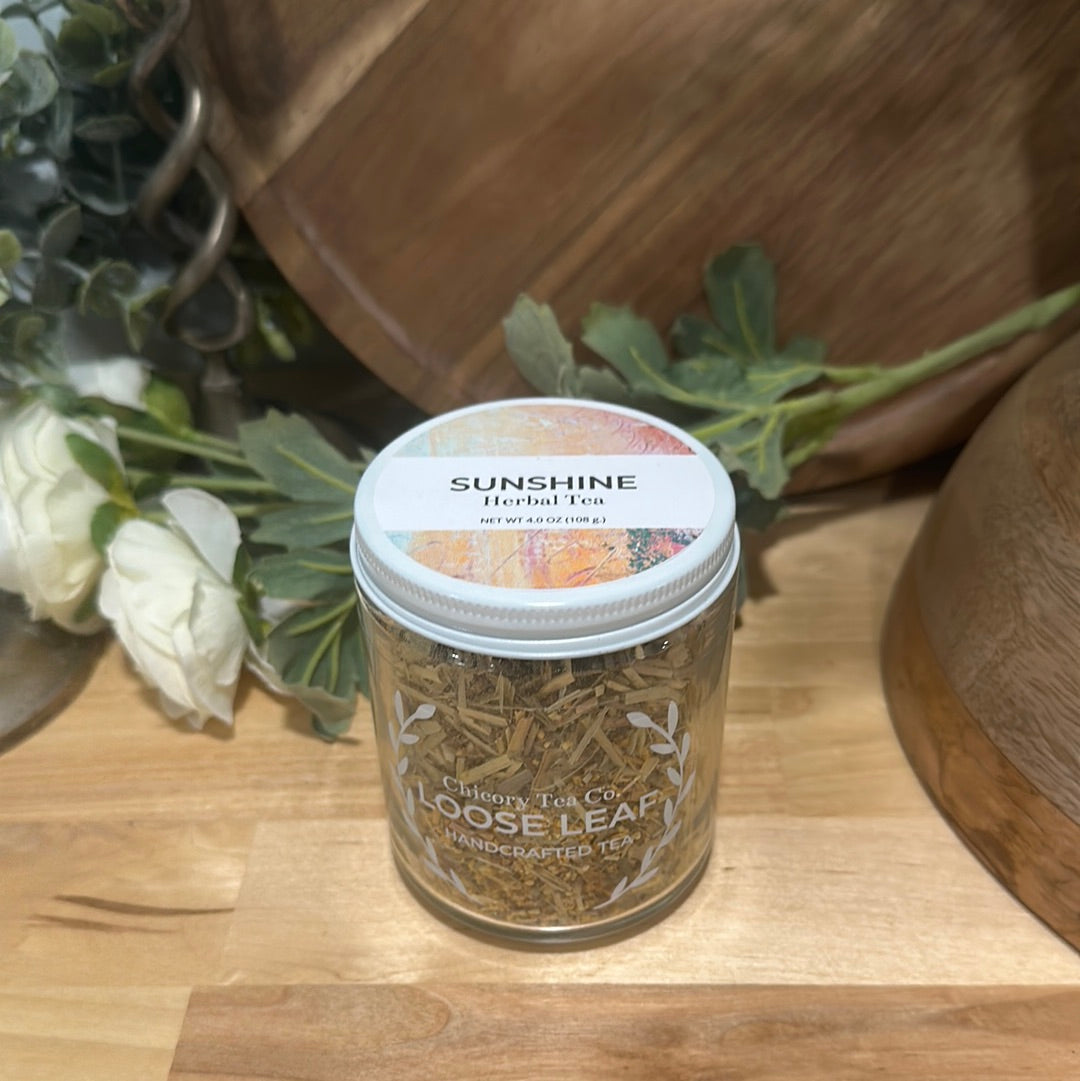 Chicory Tea Co.'s Sunshine Herbal Tea with a view from the front in the jar, on a wooden floor with a vine and a barrel in the background.