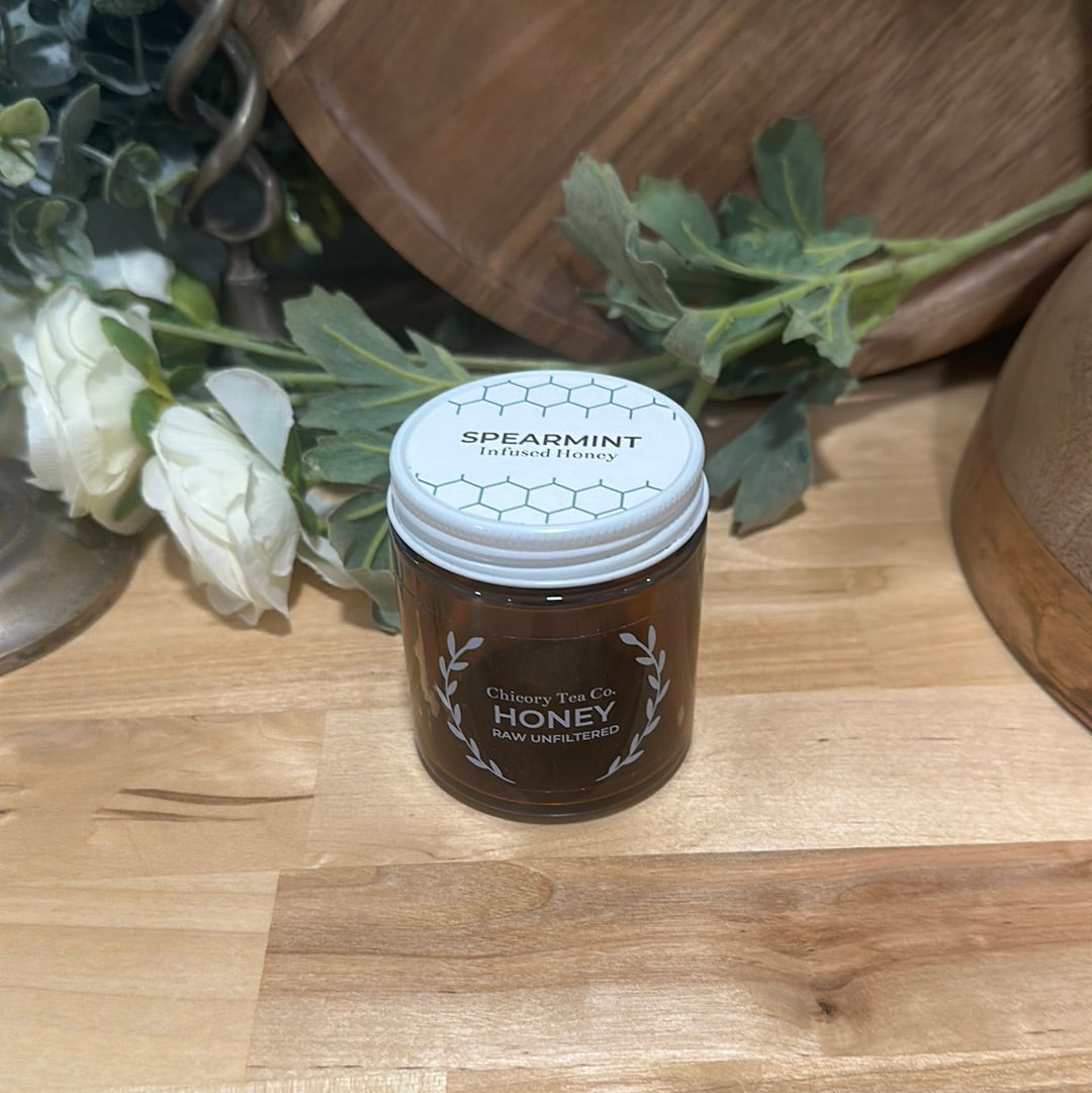 Chicory Tea Co.'s Spearmint Honey in the jar, view from front on a wood floor with flowers and a barrel in the background.