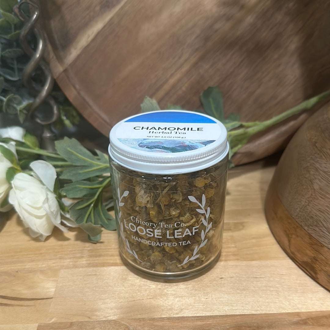 An artistic product photo of Chicory Tea Co.'s Chamomile Herbal Tea in the jar, view from front on a wood floor with flowers and barrel in the background