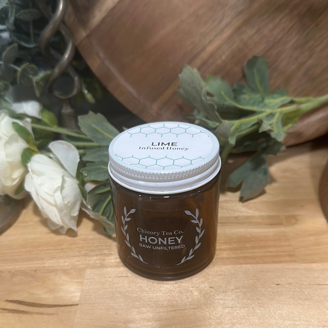 An artistic product photo of Chicory Tea Co.'s Lime Honey in the jar, view from front on a wood floor with a flowers and a barrel in the background