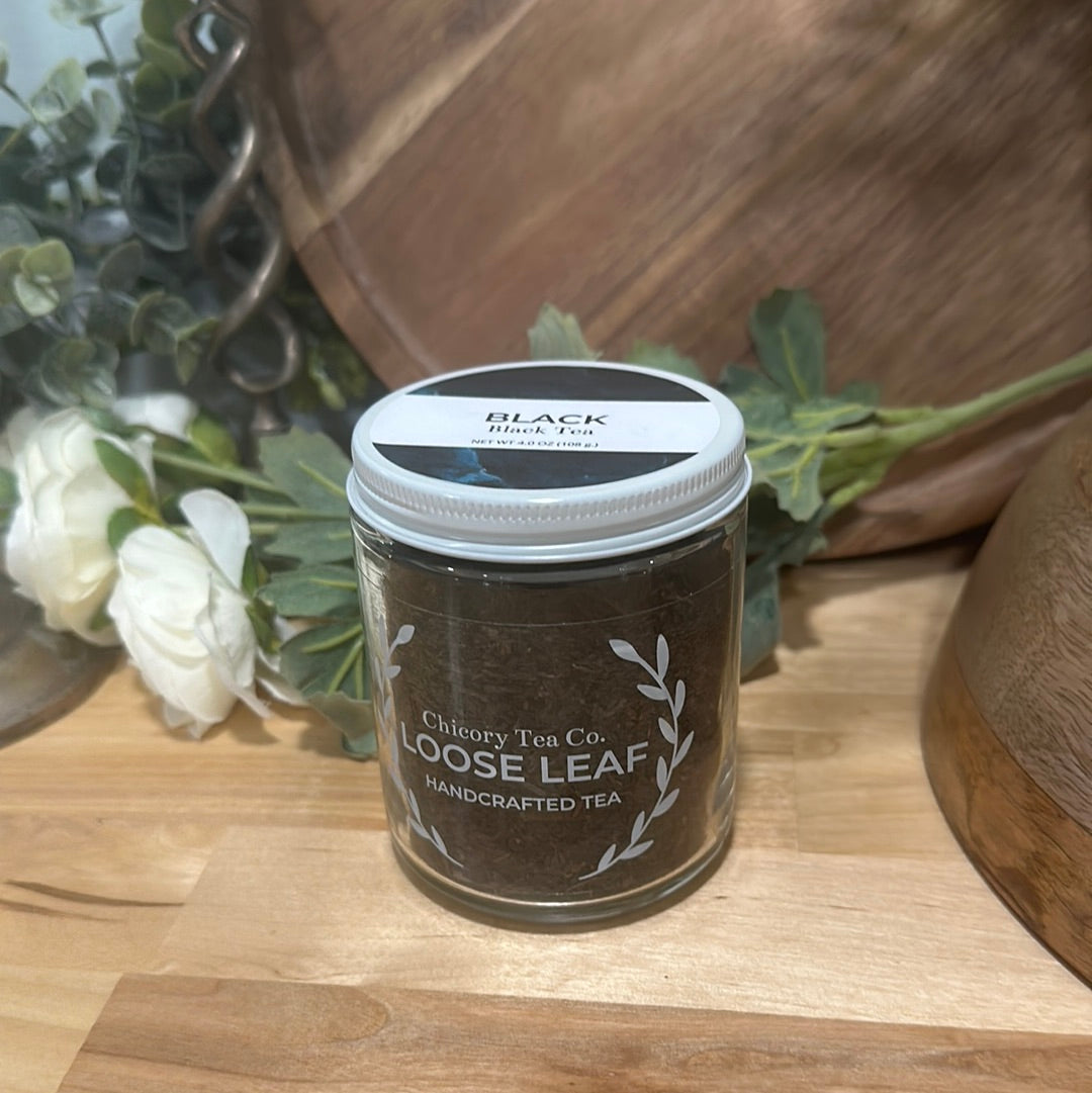 An artistic product photo of Chicory Tea Co.'s Black Tea in the jar, view from front on wood floor with flowers and barrel in the background