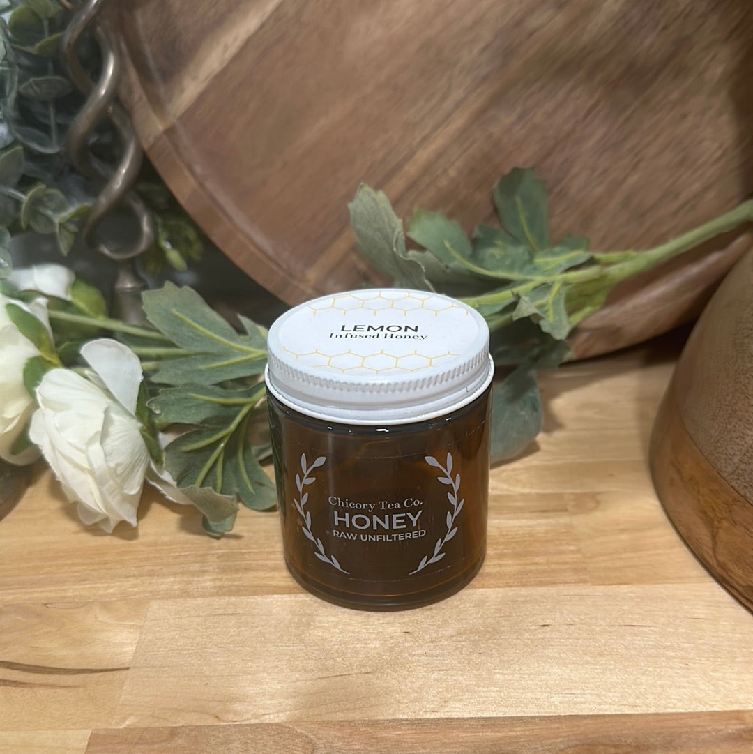 An artistic product photo of Chicory Tea Co.'s Lemon Honey in the jar, view from front on a wood floor with flowers and barrel in the background
