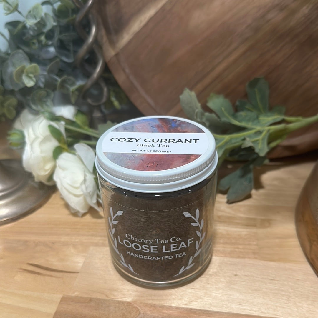 An artistic product photo of Chicory Tea Co.'s Cozy Currant in the jar, view from the front on a wood floor with flowers and barrel in the background
